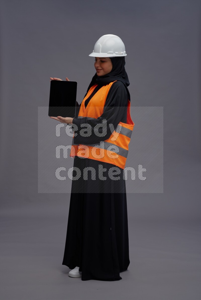 Saudi woman wearing Abaya with engineer vest standing showing tablet to camera on gray background