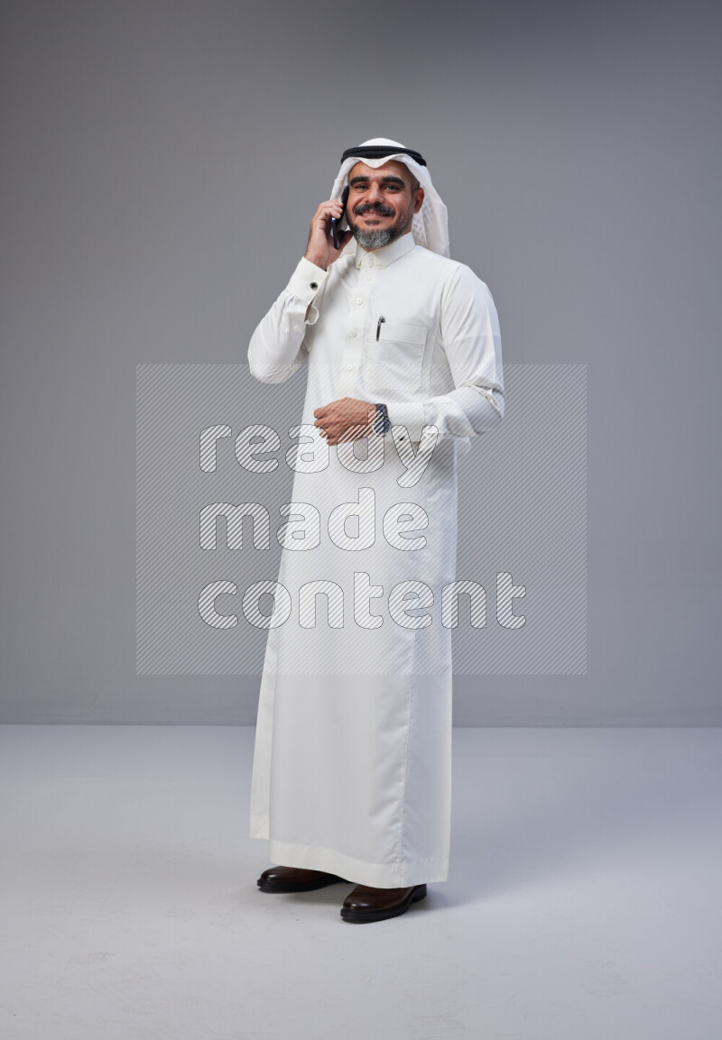 Saudi man Wearing Thob and white Shomag standing talking on phone on Gray background