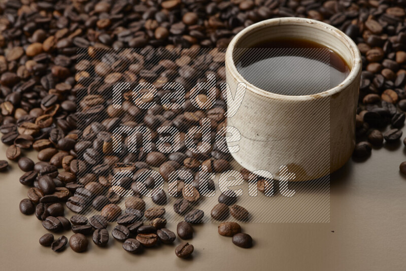 A beige pottery cup of coffee surrounded by roasted coffee beans on beige background