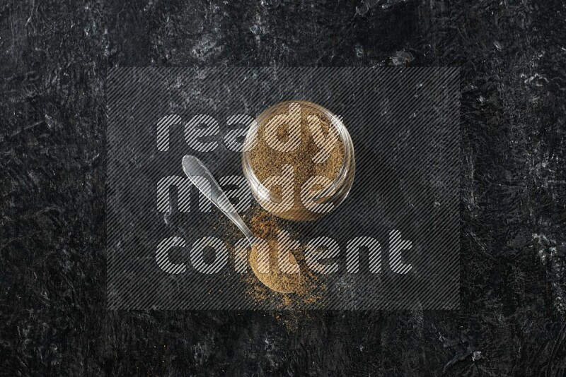 A glass jar and a metal spoon full of cumin powder on a textured black flooring