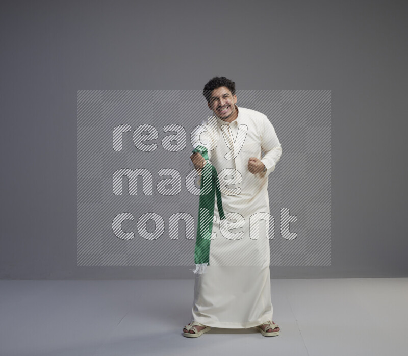 A Saudi man standing wearing thob holding Saudi flag scarf on gray background