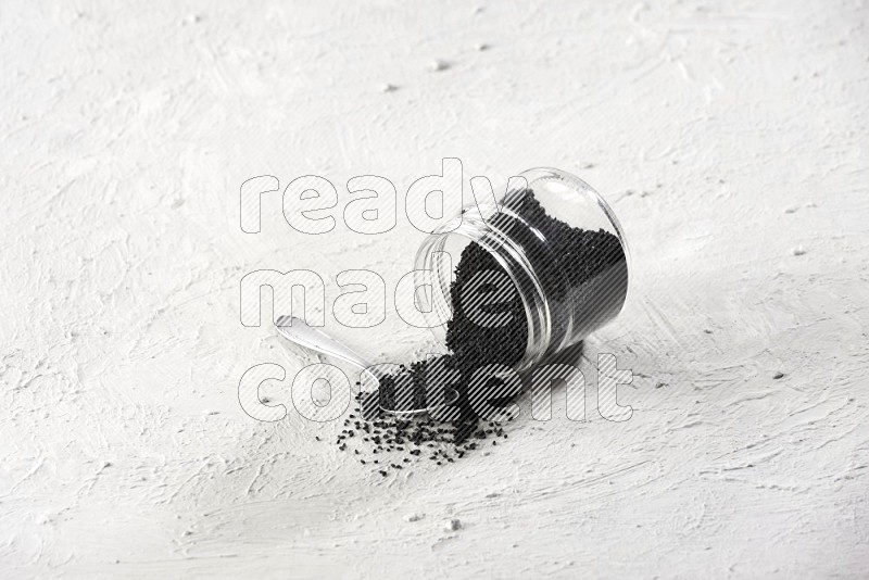 A glass jar and a metal spoon full of black seeds and the jar flipped and seeds spread on a textured white flooring