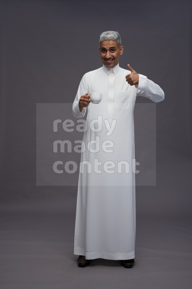 Saudi man wearing thob standing holding mug on gray background