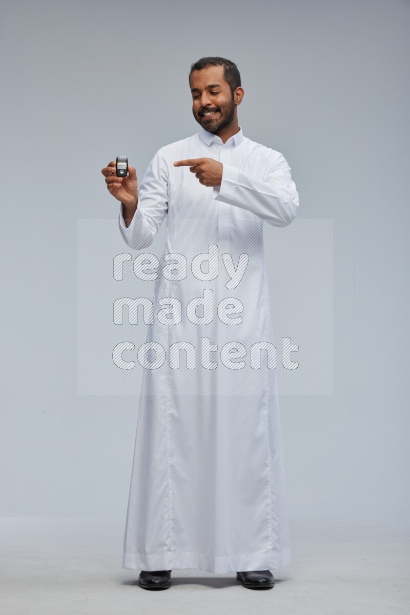 Saudi man wearing thob and shomag standing holding car key on gray background