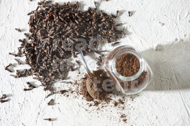 A glass spice jar and a metal spoon full of cloves powder and cloves spread on textured white flooring
