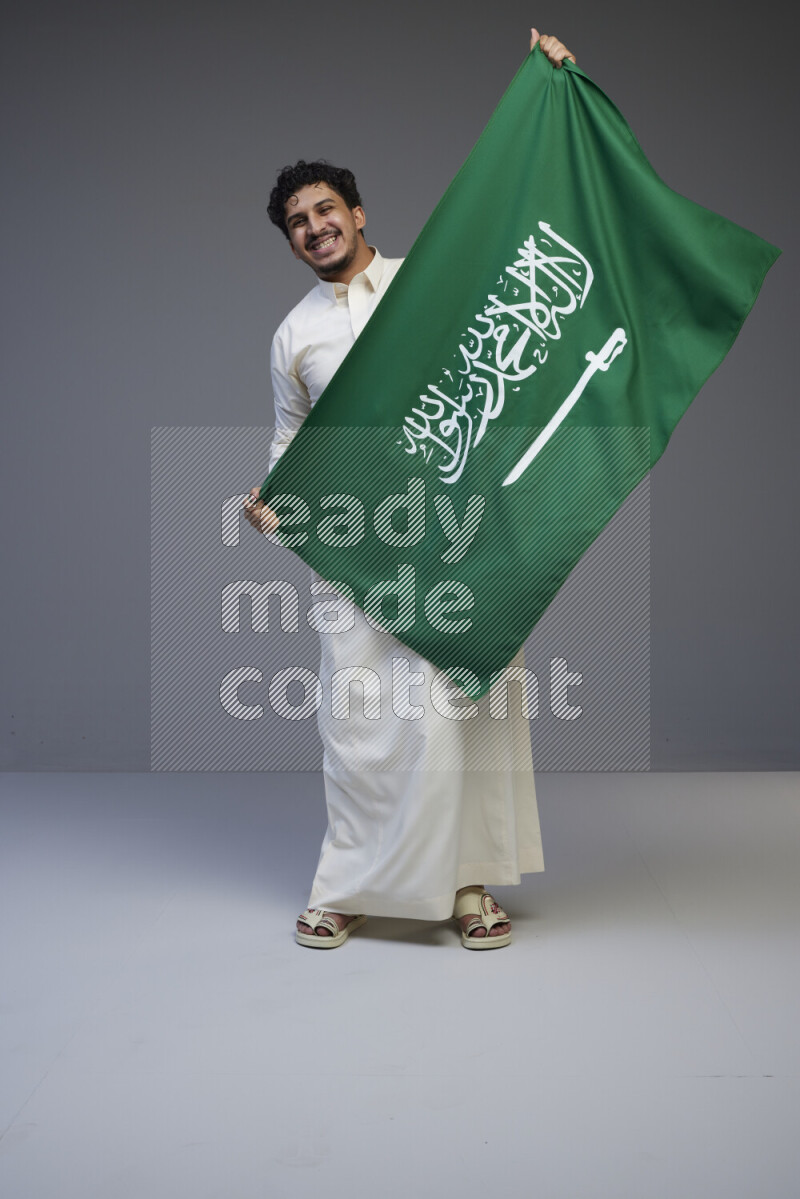 A Saudi man standing wearing thob holding big Saudi flag on gray background