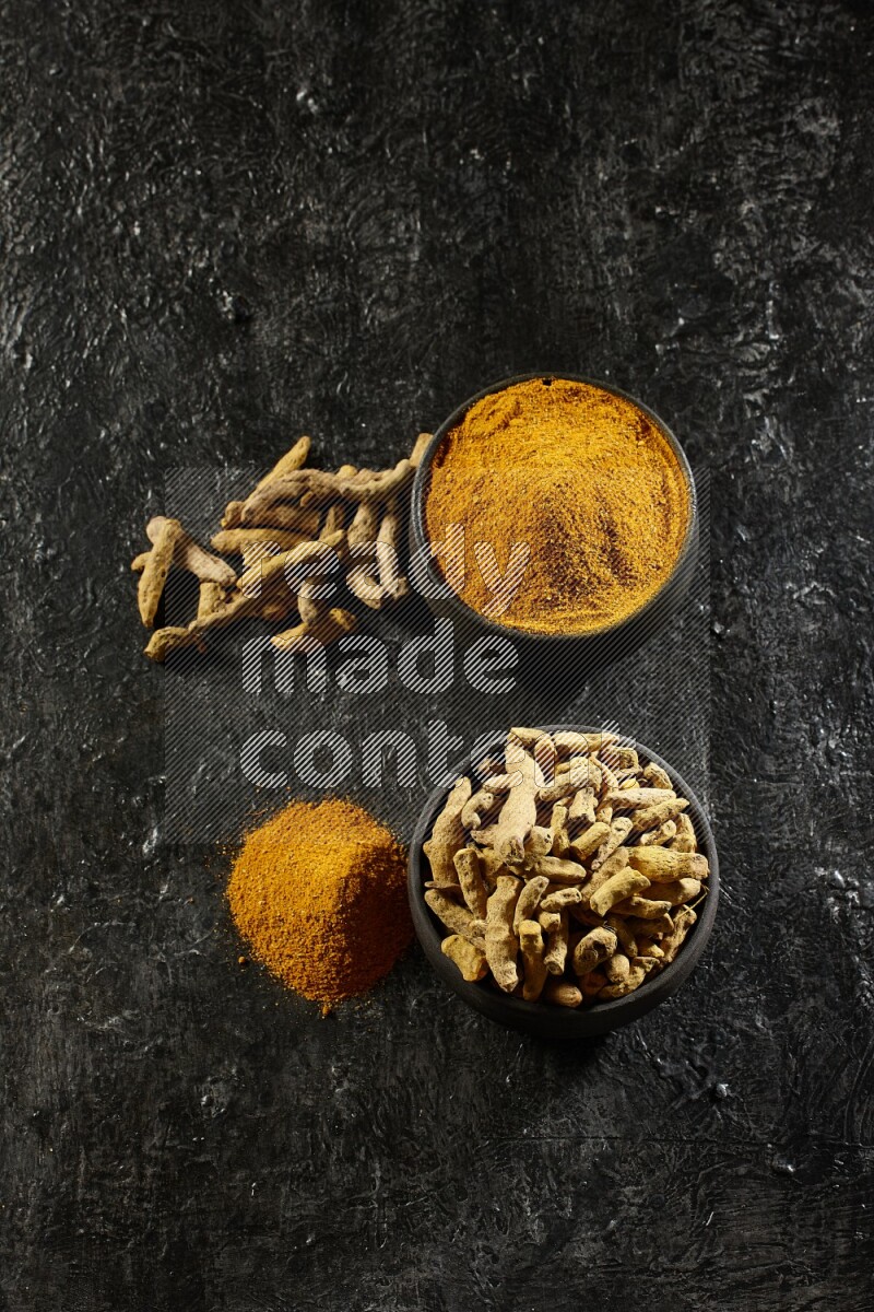 2 pottery black bowls, one full of turmeric powder and the other full of dried turmeric whole fingers on a textured black flooring