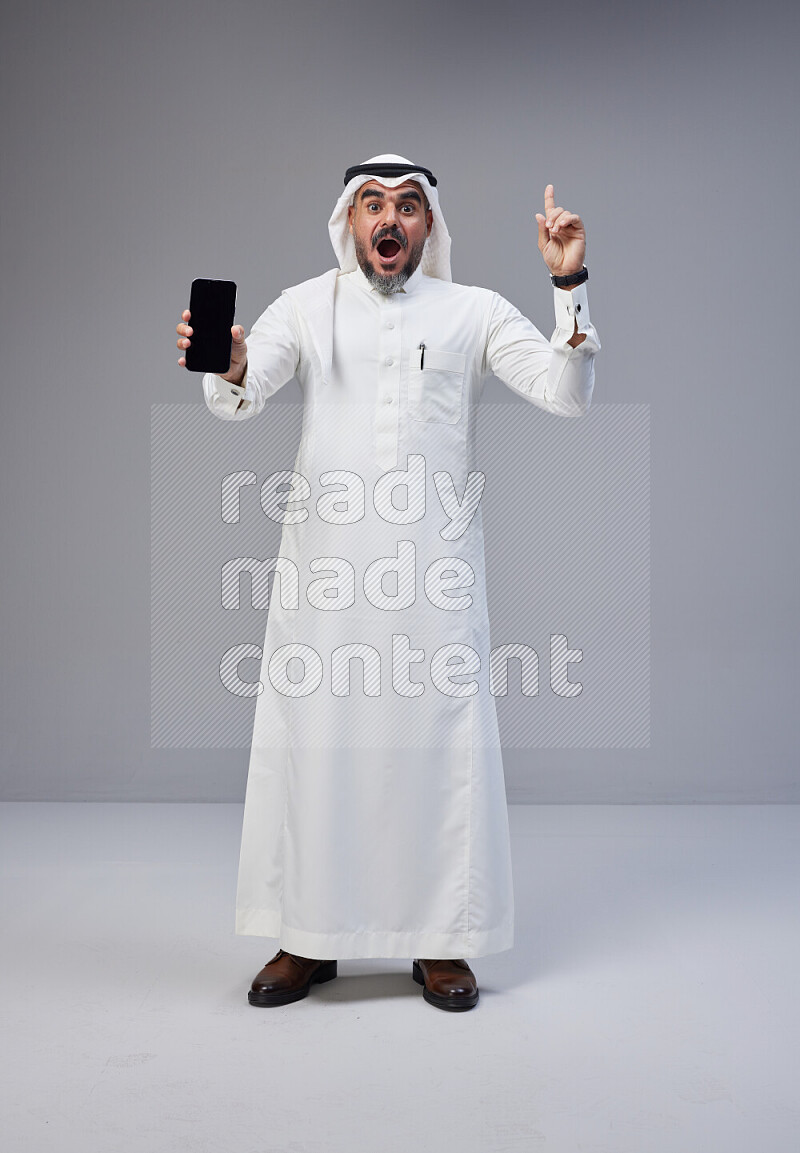 Saudi man Wearing Thob and white Shomag standing showing phone to camera on Gray background