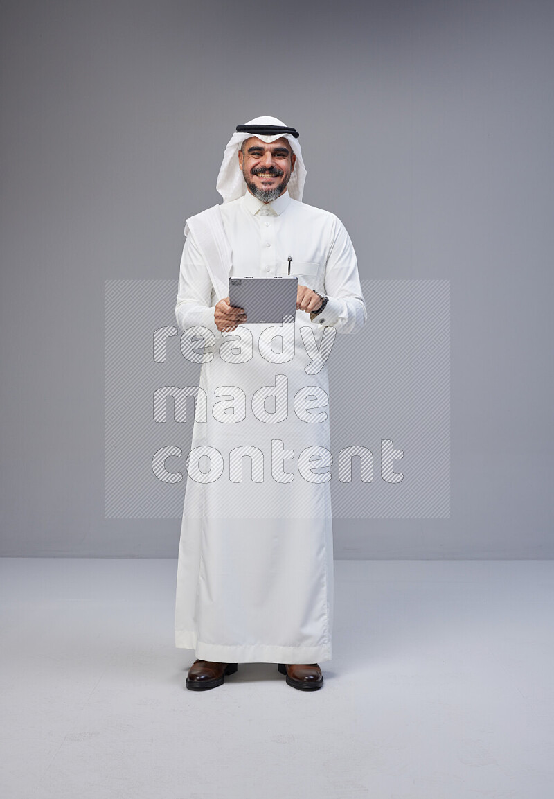 Saudi man Wearing Thob and white Shomag standing working on tablet on Gray background