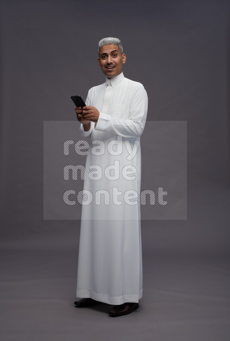 Saudi man wearing thob standing texting on phone on gray background