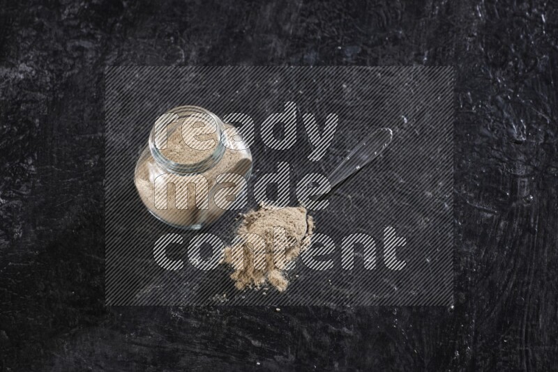 A glass spice jar full of garlic powder with a filled metal spoon on a textured black flooring