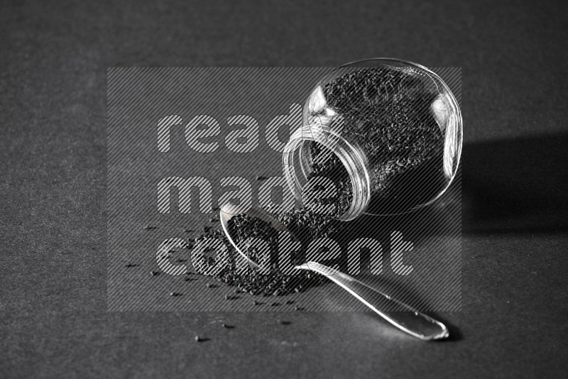 وعاء بهارات زجاجي مقلوب ممتلئ ببذور حبة البركة والبذور تناثرت منه مع ملعقة معدنية ممتلئة على أرضية سوداء