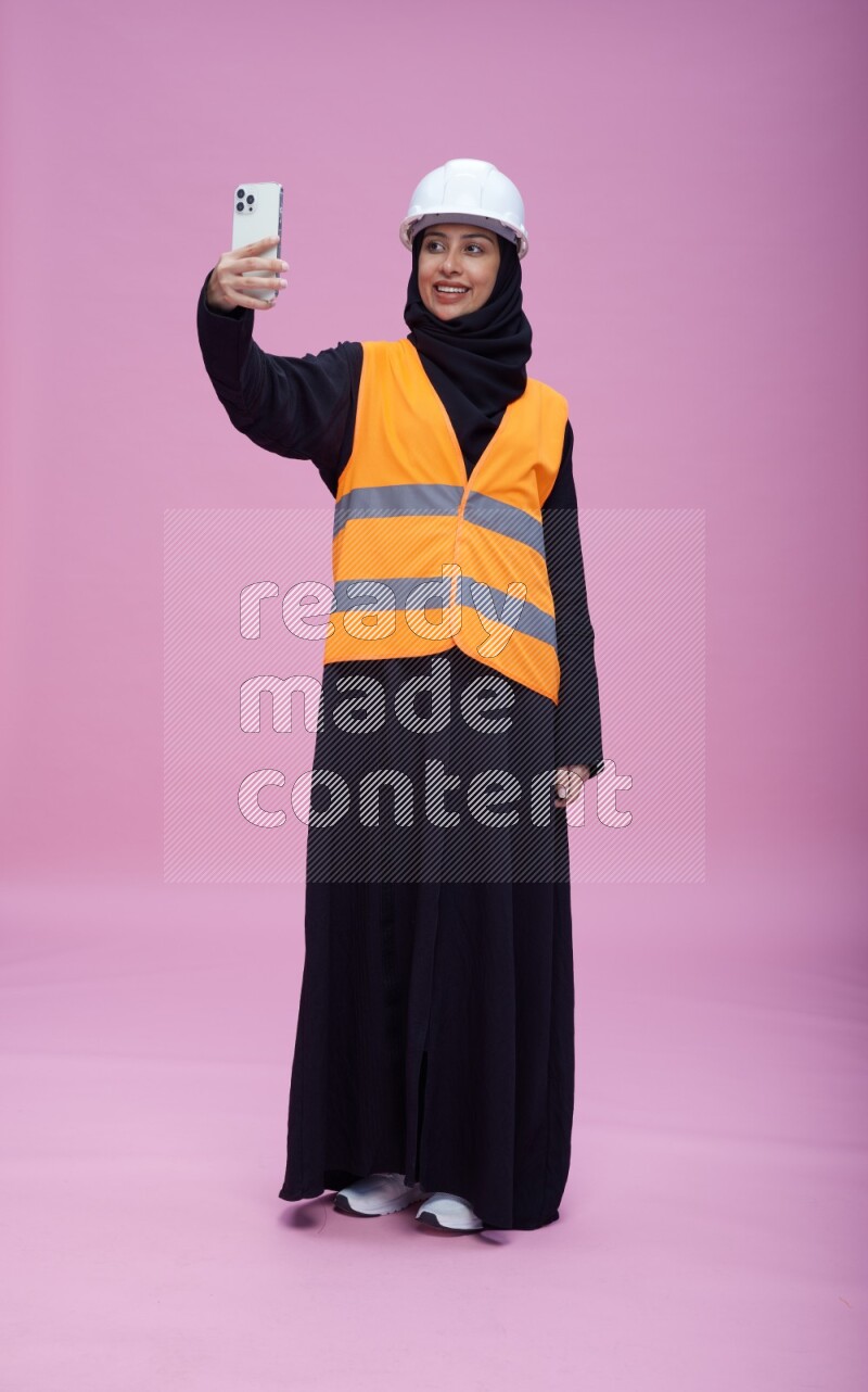 Saudi woman wearing Abaya with engineer vest and helmet standing taking selfie on pink background