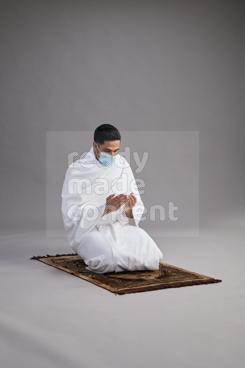 A man wearing Ehram with face mask sitting on floor performing dua'a on gray background