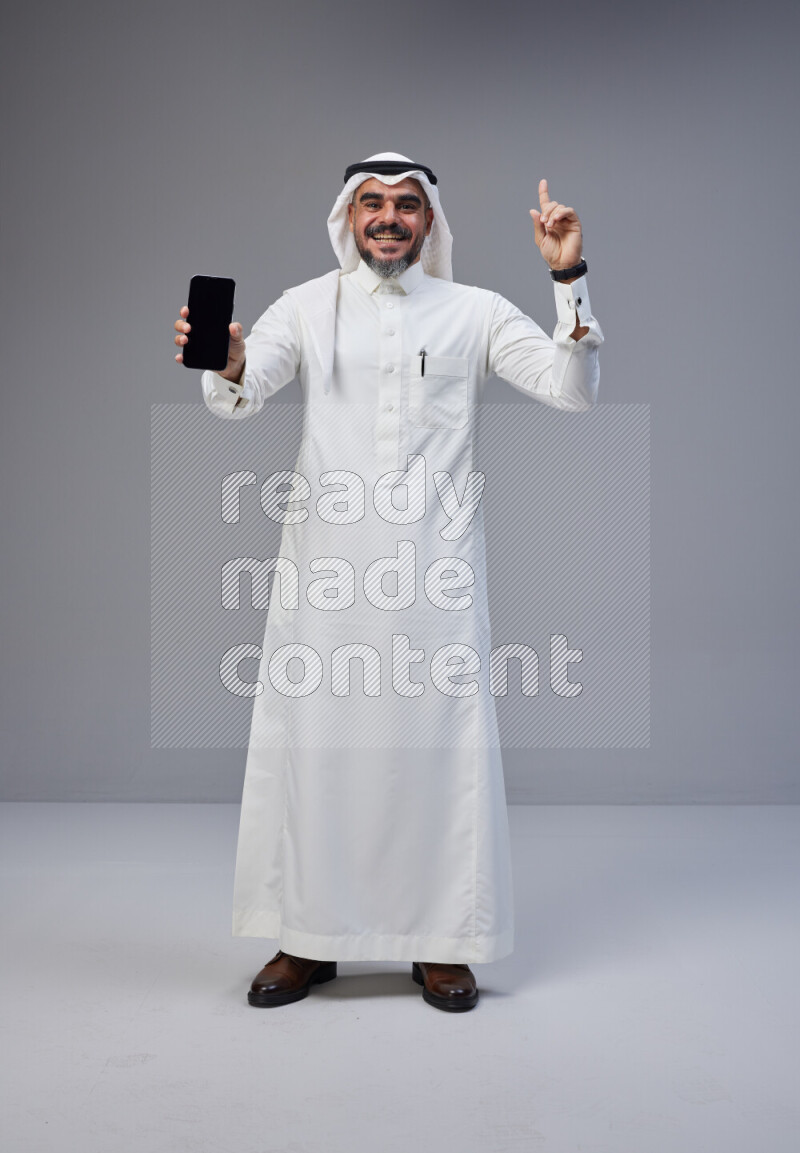 Saudi man Wearing Thob and white Shomag standing showing phone to camera on Gray background