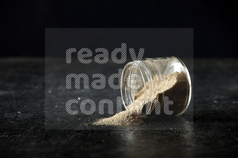 A flipped glass jar full of cardamom powder and powder spilled out of it on textured black flooring