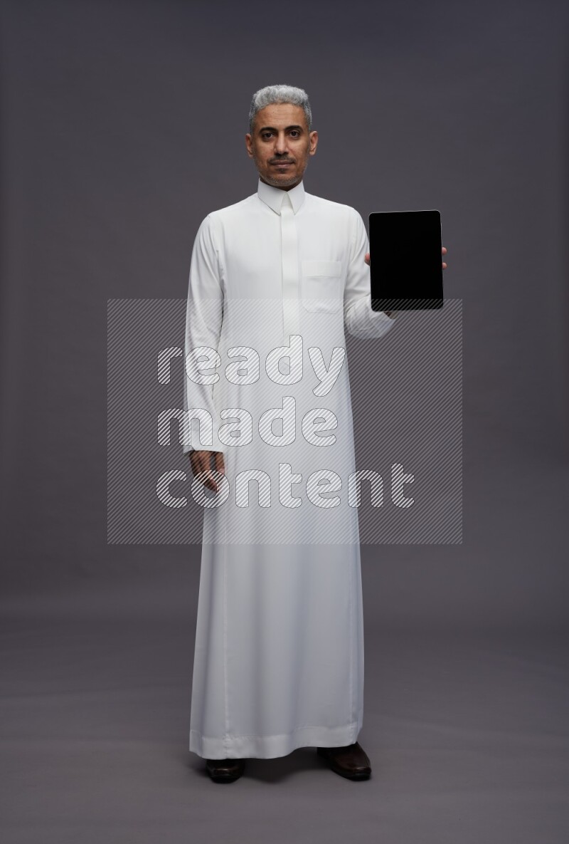 Saudi man wearing thob standing showing tablet to camera on gray background