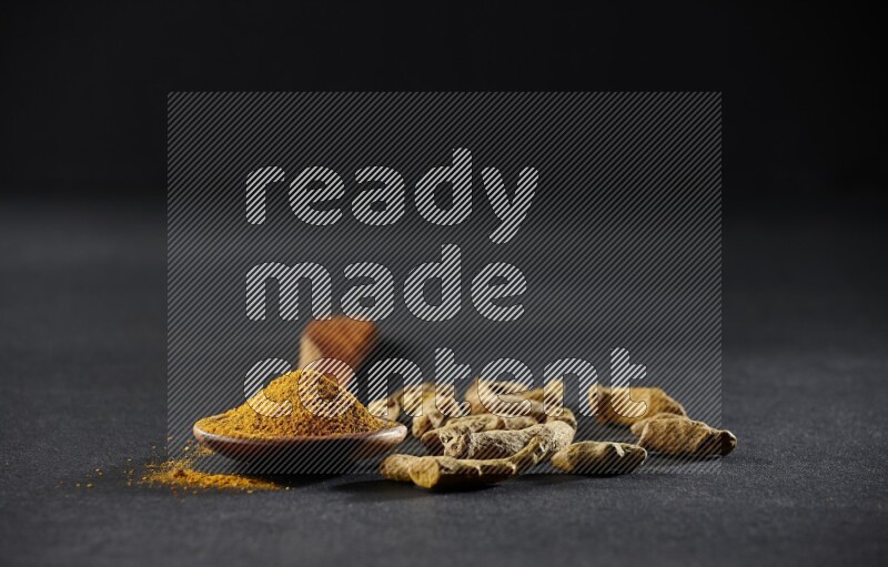 مغرفة خشبية ممتلئة ببودرة الكركم مع أصابع الكركم المجففة على خلفية سوداء