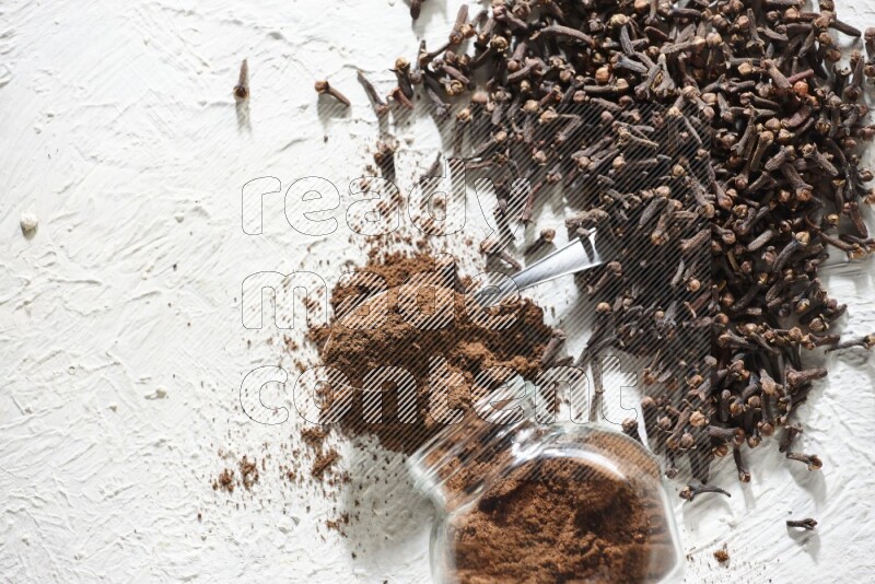 A flipped glass spice jar and a metal spoon full of cloves powder and powder came out of the jar with cloves spread on textured white flooring