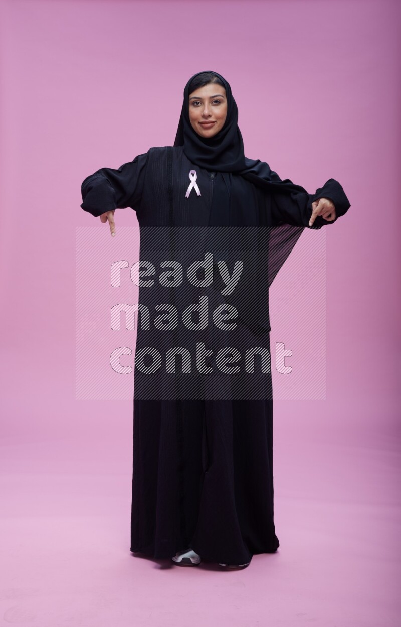 Saudi woman wearing pink ribbon on Abaya standing interacting with the camera on pink background
