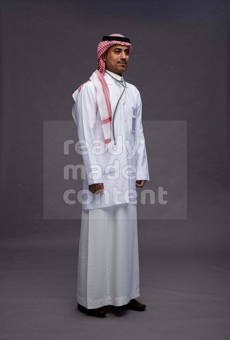 Saudi man wearing thob with lab coat and shomag with pocket employee badge with stethoscope standing interacting with the camera on gray background