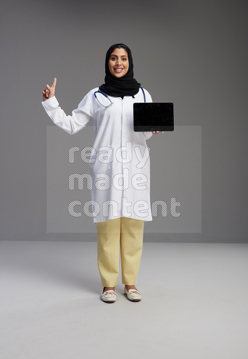 Saudi woman wearing lab coat with stethoscope standing showing tablet to camera with sign in the back on Gray background