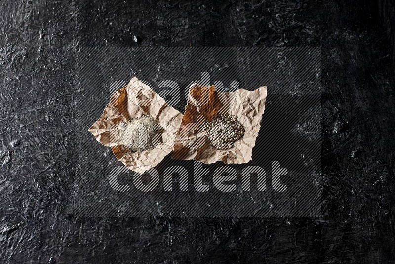 White pepper beads and powder in 2 crumpled paper on textured black flooring