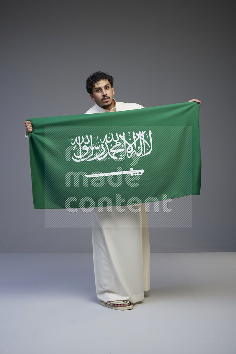 A Saudi man standing wearing thob holding big Saudi flag on gray background