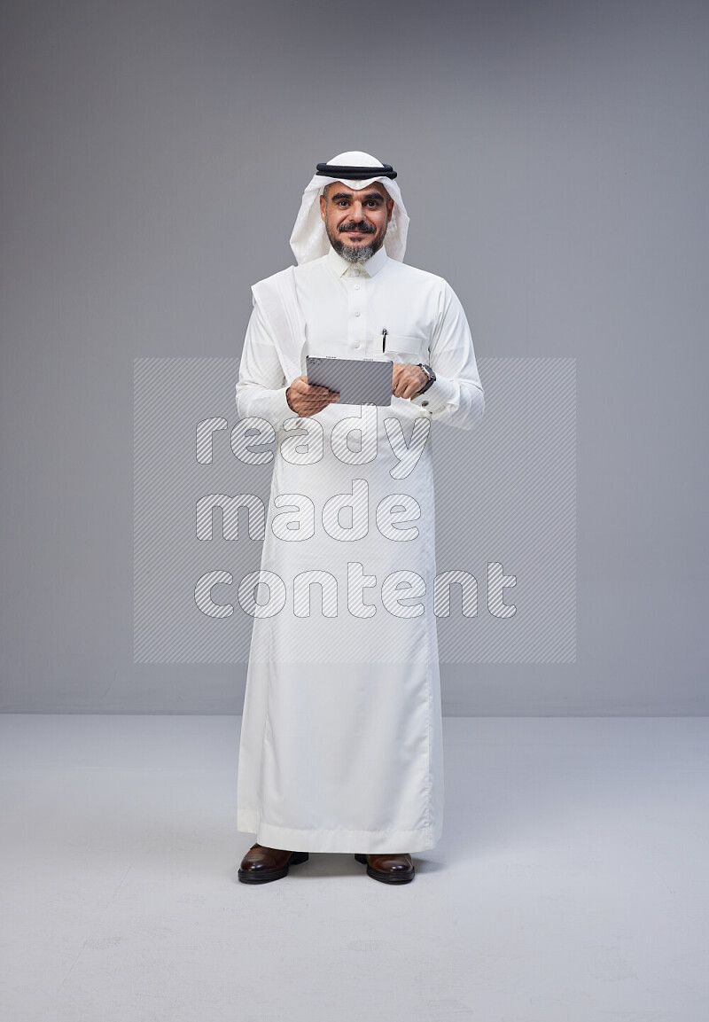 Saudi man Wearing Thob and white Shomag standing working on tablet on Gray background