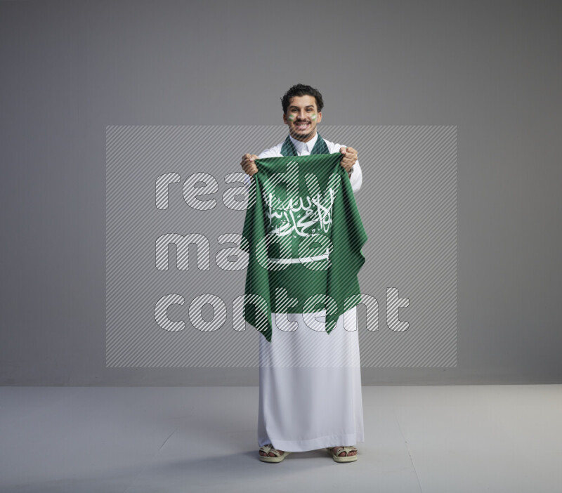 A Saudi man standing wearing thob with face painting wrapping Saudi flag scarf and holding big Saudi flag on gray background