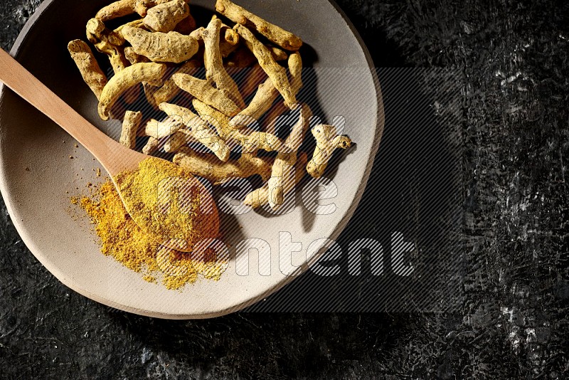 A plate filled with dried turmeric fingers and a wooden spoon full of turmeric powder on a textured black flooring