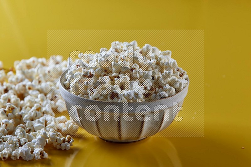 A white pottery bowl full of popcorn with popcorn beside it on a yellow background in different angles