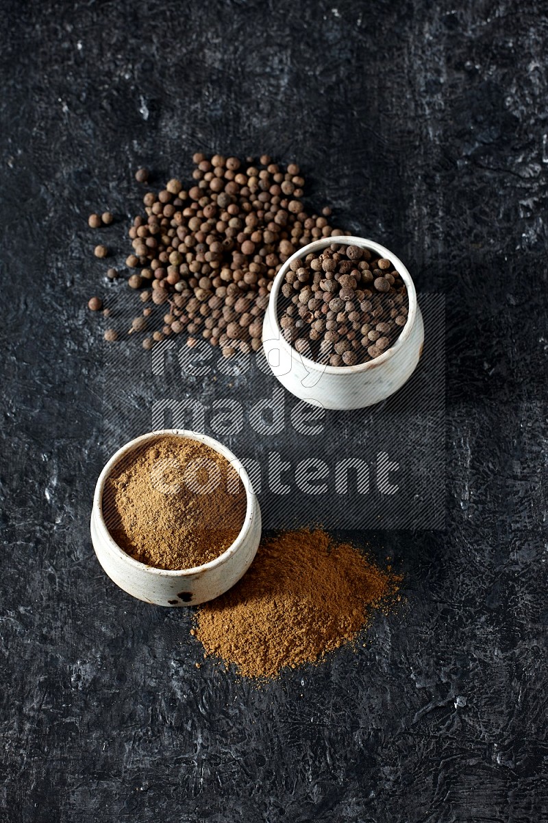 2 beige bowls, one full of allspice powder and the other full of whole balls and both spread on a textured black flooring