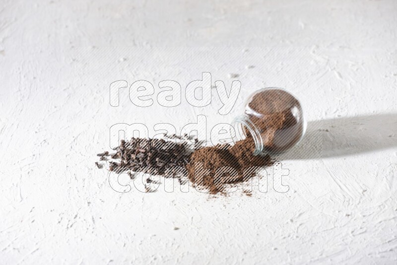 A glass spice jar full of cloves powder flipped and powder came out of it with cloves spread on textured white flooring