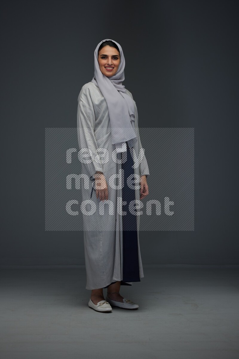 A Saudi woman wearing a light gray Abaya and head scarf standing and making multi poses eye level on a grey background