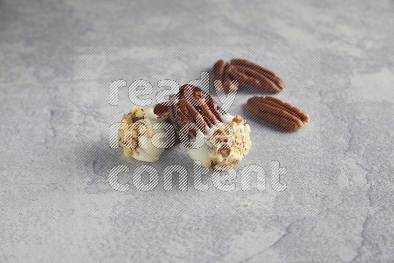 Two pecan stuffed dates covered with white chocolate and chopped walnuts with unroasted pecans on alight grey background