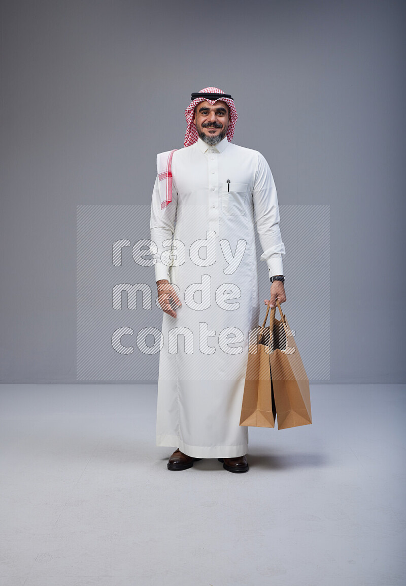 Saudi man Wearing Thob and red Shomag standing holding shopping bag on Gray background