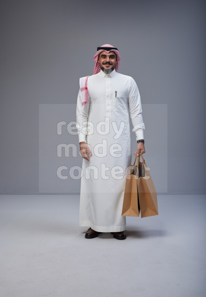 Saudi man Wearing Thob and red Shomag standing holding shopping bag on Gray background