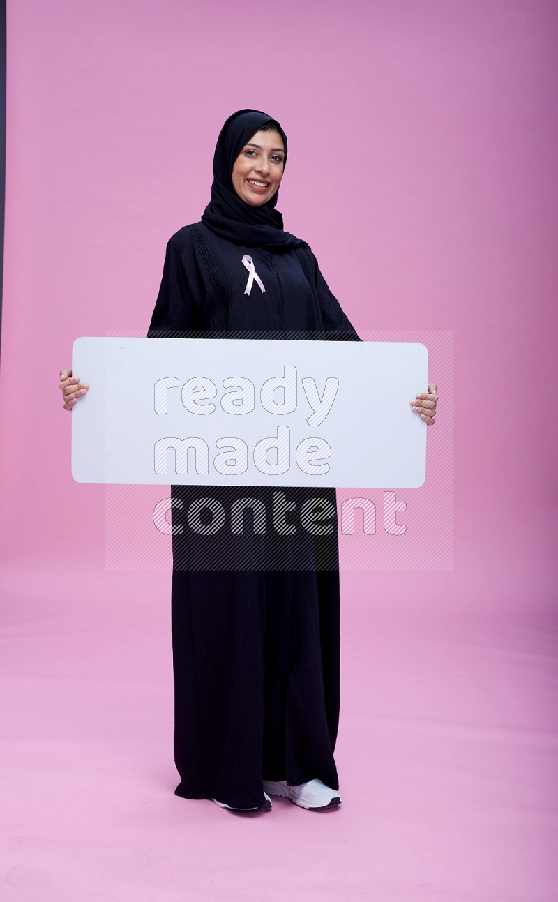 Saudi woman wearing pink ribbon on Abaya standing holding board on pink background