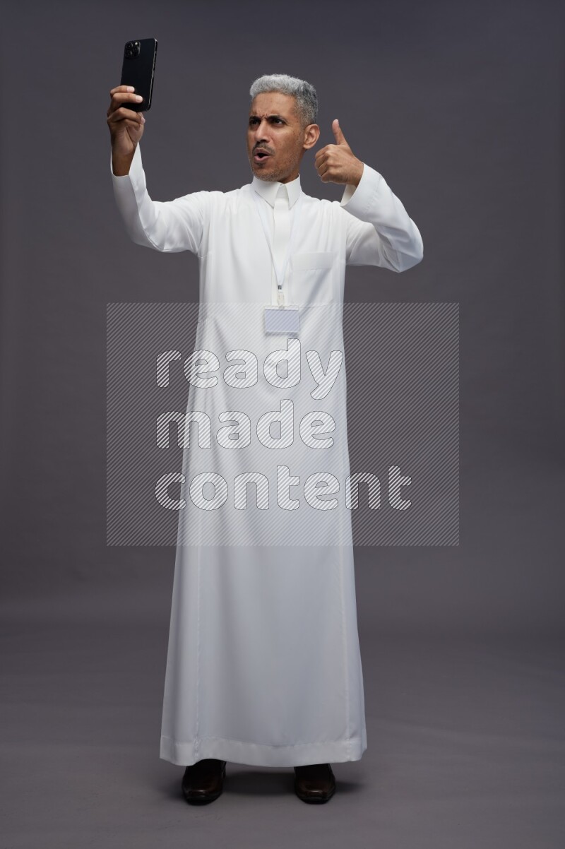 Saudi man wearing thob with neck strap employee badge standing taking selife on gray background