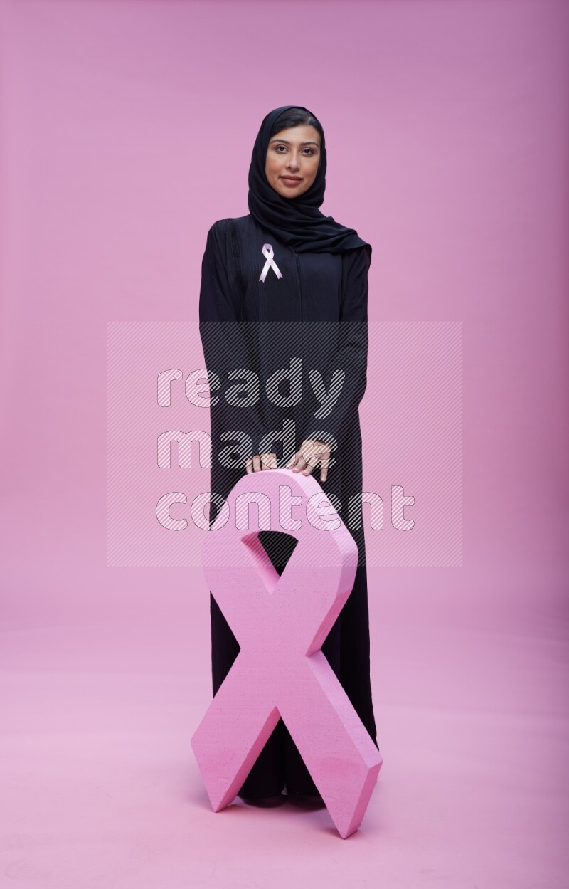 Saudi woman wearing pink ribbon on abaya standing holding awareness ribbon on plain pink background