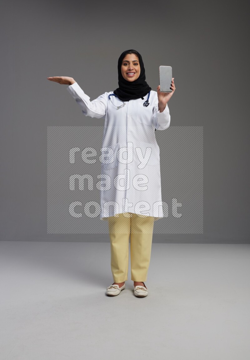 Saudi woman wearing lab coat with stethoscope standing showing phone to camera with sign in the back on Gray background
