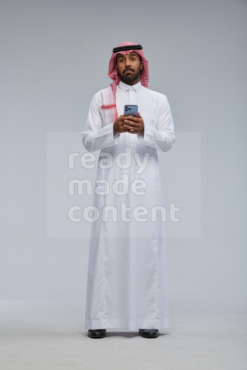 Saudi man Wearing Thob and shomag standing texting on phone on Gray background