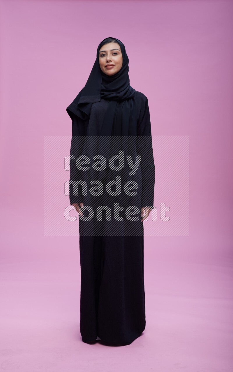 Saudi woman wearing Abaya standing interacting with the camera on pink background
