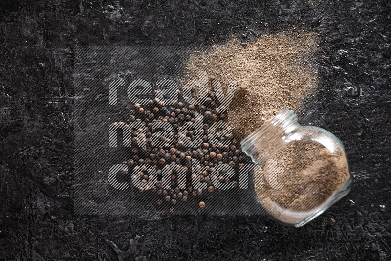 A flipped glass spice jar full of black pepper powder with pepper beads on textured black flooring