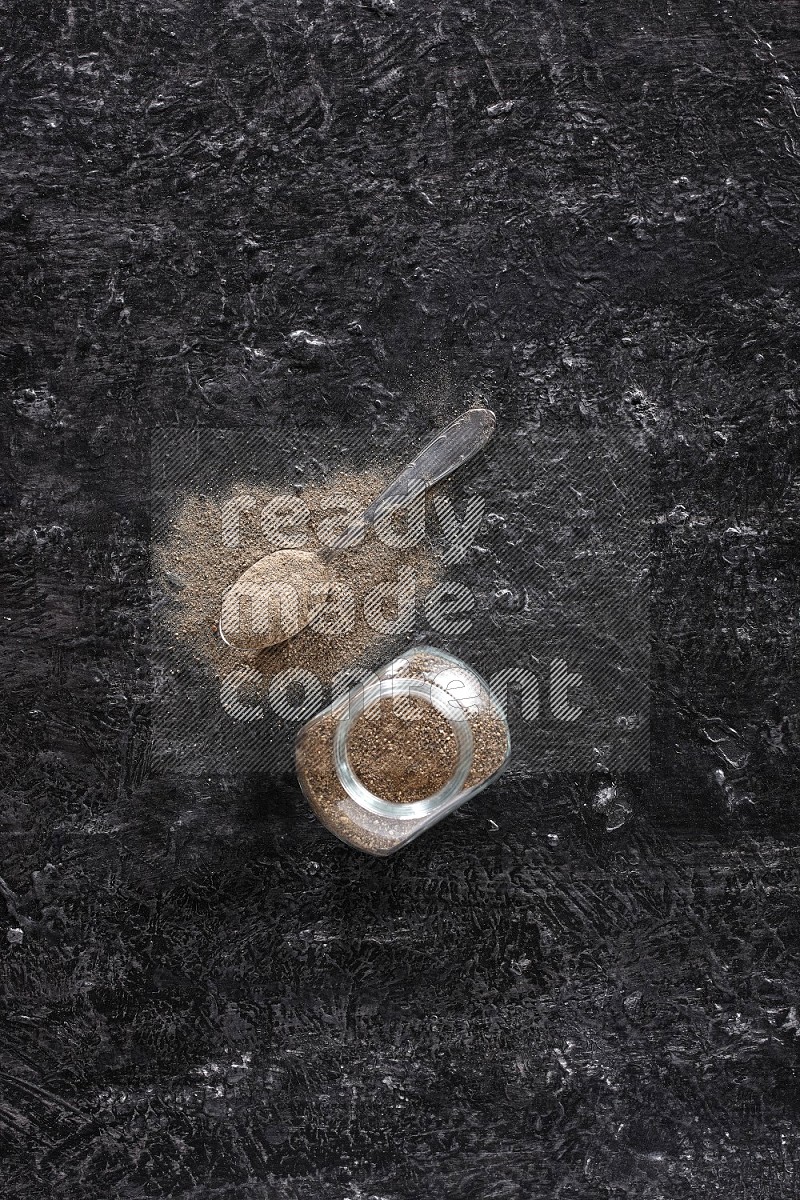 A glass spice jar full of black pepper powder and a metal spoon full of powder on textured black flooring