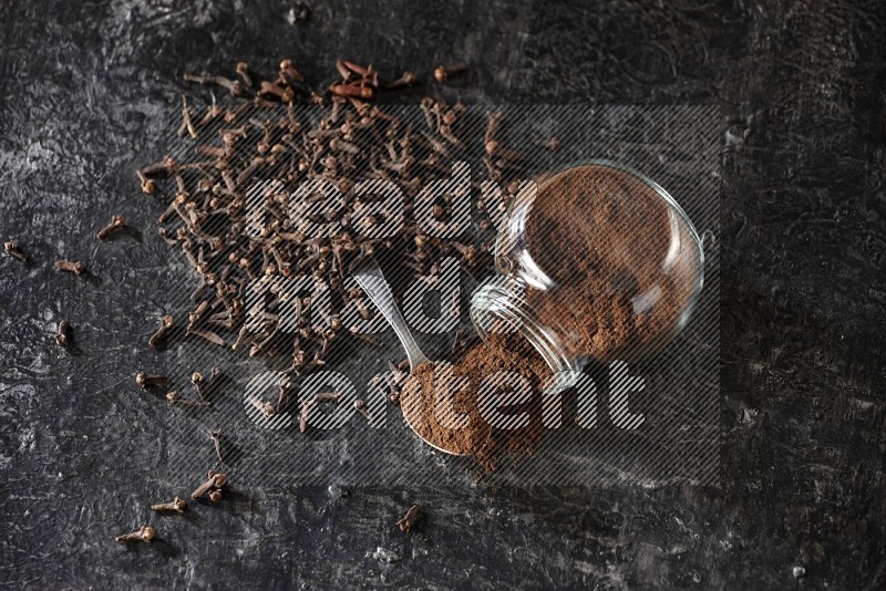 A flipped glass spice jar and a metal spoon full of cloves powder with cloves spread on textured black flooring