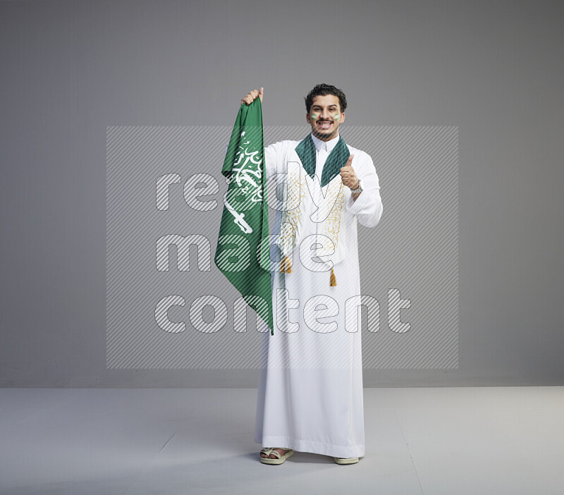 A Saudi man standing wearing thob with face painting wrapping Saudi flag scarf and holding big Saudi flag on gray background