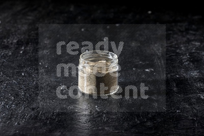 A glass jar full of white pepper powder on textured black flooring