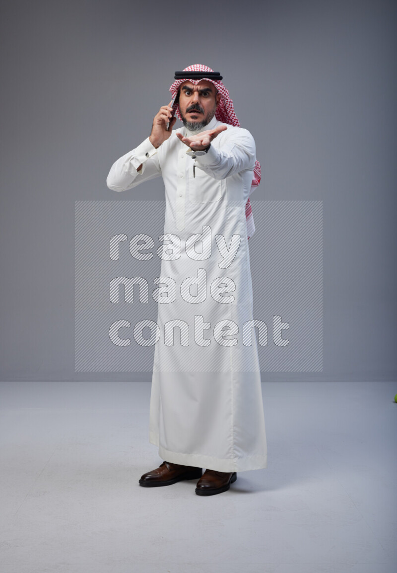 Saudi man Wearing Thob and red Shomag standing talking on phone on Gray background